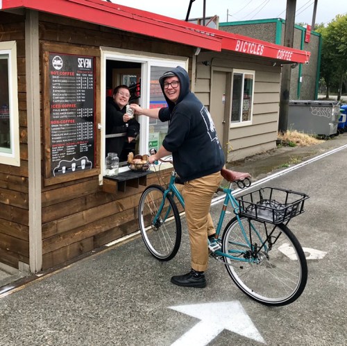 A person on a bicycle receives coffee and pastries from a smiling barista at a small outdoor coffee stand.