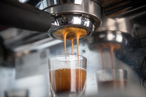 Close-up of espresso being brewed, with rich coffee flowing into a glass shot glass. Reflective metal background.