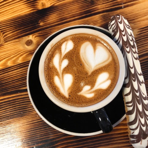 A cup of coffee with heart and leaf latte art, accompanied by a striped chocolate biscuit on a wooden table.