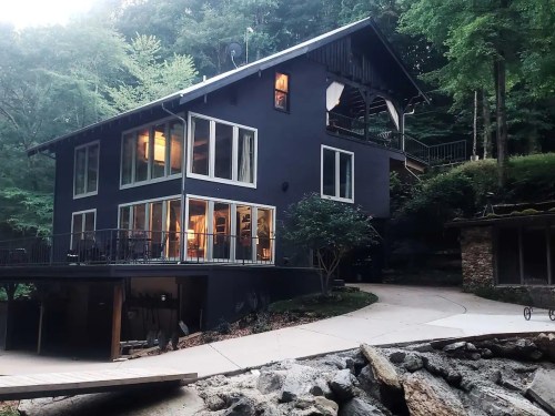 A modern black house with large windows, surrounded by trees, featuring a curved driveway and outdoor seating area.