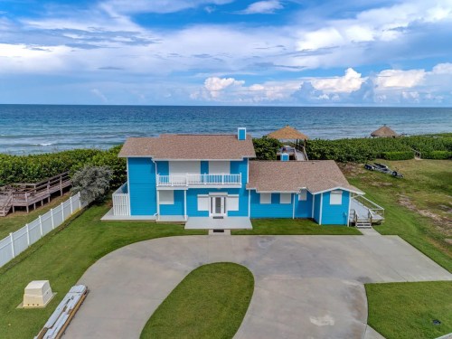 A bright blue beach house with a large lawn, overlooking the ocean under a partly cloudy sky.