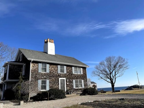 A two-story house with shingle siding, a chimney, and a view of the ocean under a clear blue sky.