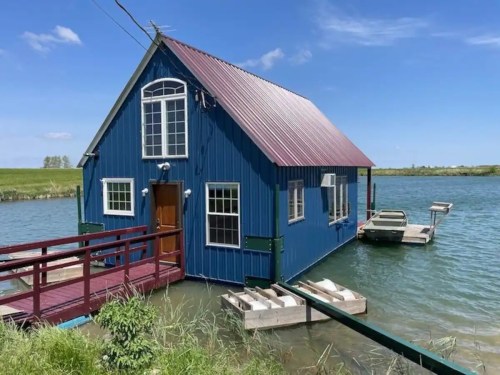 A blue house with a red roof on stilts over water, featuring a small dock and surrounded by greenery under a clear sky.