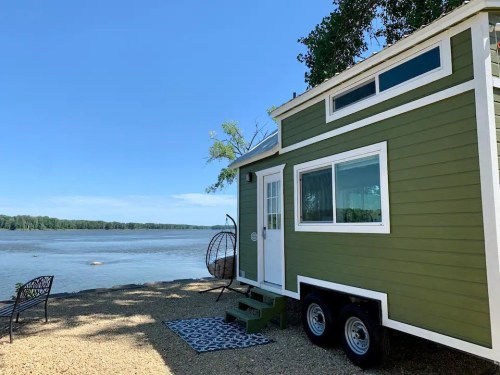 A green tiny house on wheels by a river, with a small porch and a hanging chair nearby, under a clear blue sky.
