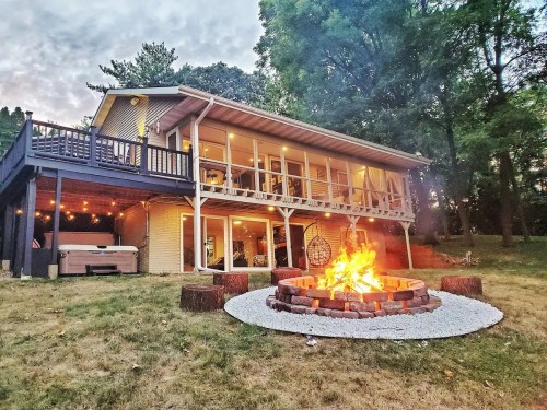 A cozy house with a deck overlooks a fire pit surrounded by logs, set in a grassy area with trees in the background.