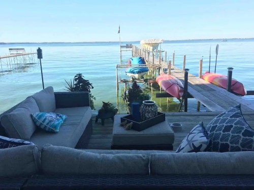 A cozy outdoor seating area overlooks a calm lake with a wooden dock and colorful kayaks in the background.