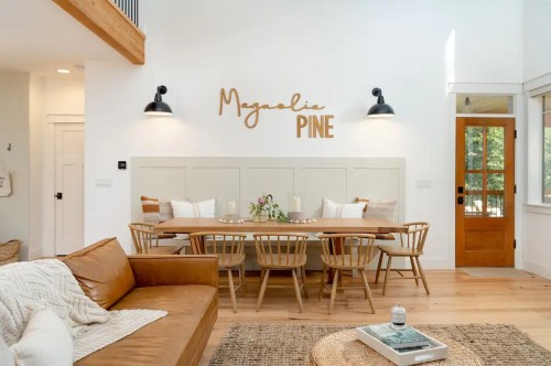 A bright dining area with a long wooden table, chairs, and a "Magnolia Pine" sign on the wall. Cozy decor elements included.