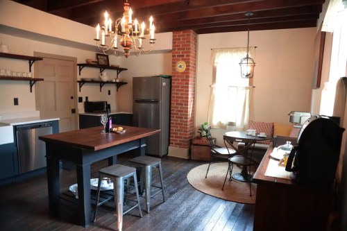 Cozy kitchen with wooden beams, a chandelier, dining table, and modern appliances against a brick wall.
