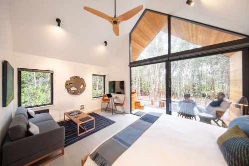 A modern, airy room with a bed, sofa, and large windows overlooking a forested area. Two people relax outside.