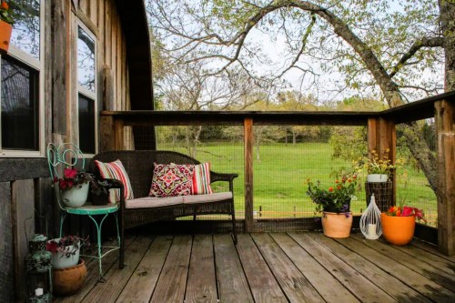 Cozy outdoor deck with a wicker bench, colorful cushions, and potted plants, overlooking a green landscape.