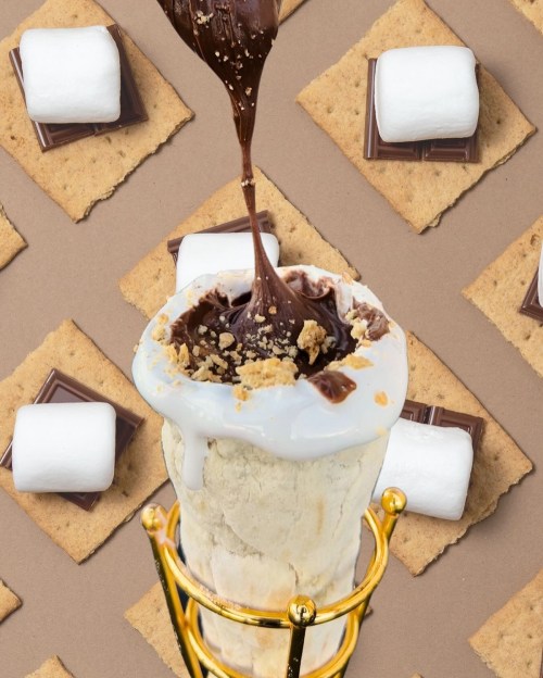 A dessert cup filled with ice cream, topped with chocolate drizzle, surrounded by graham crackers and marshmallows.