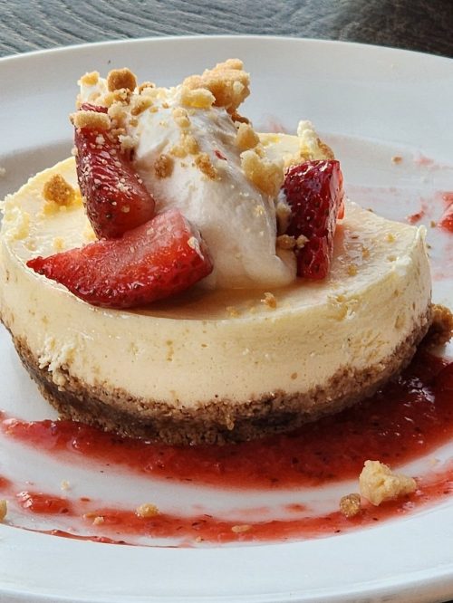 A slice of cheesecake topped with strawberries, whipped cream, and crumbs, served on a white plate with strawberry sauce.