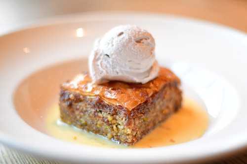 A square piece of baklava topped with a scoop of ice cream, served on a white plate with a drizzle of syrup.