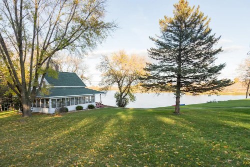 A serene lakeside view featuring a house, trees, and a grassy area with autumn leaves scattered on the ground.