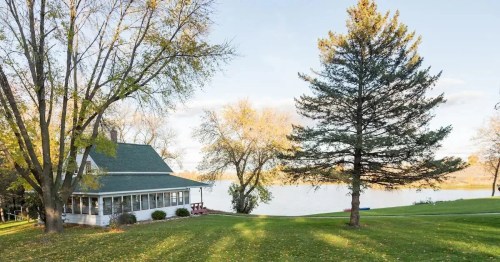 A serene lakeside view featuring a house, trees, and a grassy area with autumn leaves scattered on the ground.