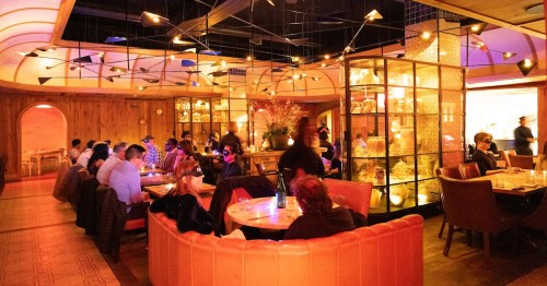 A vibrant restaurant interior with dim lighting, featuring patrons dining at round tables and a decorative glass display.