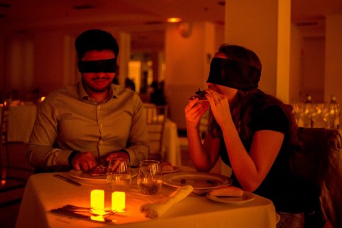 A couple dining in a dimly lit restaurant, both wearing blindfolds, with candles on the table.