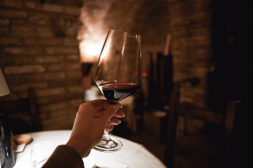 A hand holding a glass of red wine in a dimly lit, rustic restaurant setting.