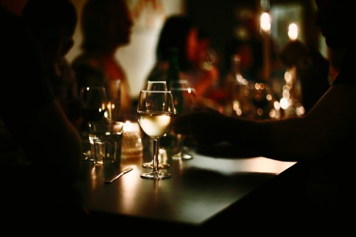 A dimly lit table with glasses of wine and water, surrounded by blurred figures enjoying a social gathering.