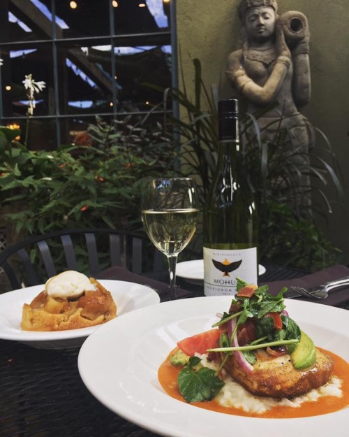 A plate of gourmet food with a glass of wine, a dessert, and a decorative statue in a lush outdoor setting.