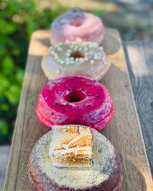 A wooden platter holds four colorful donuts: pink, beige, and chocolate, topped with sprinkles and a square of marshmallow.
