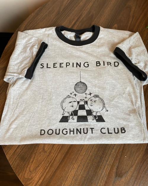 A gray t-shirt with black sleeves featuring a graphic of dancing doughnuts and the text "Sleeping Bird Doughnut Club."