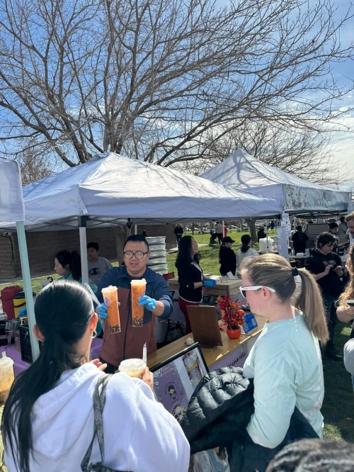 A vendor holds two drinks at a busy outdoor market, with customers and tents in the background under a clear sky.