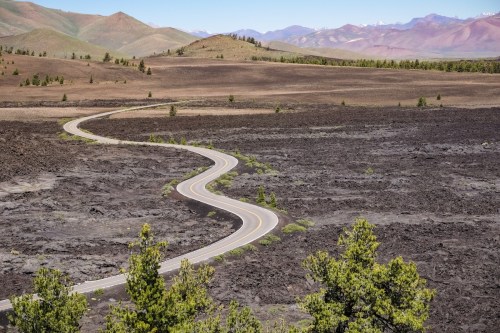 A winding road cuts through a barren landscape of dark lava rock, with distant mountains and sparse greenery.