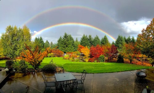 A vibrant double rainbow arcs over a colorful autumn garden with lush trees and a patio table.