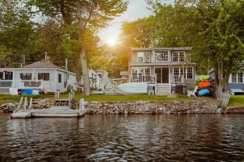A lakeside view of a house with kayaks, surrounded by trees and a sunset reflecting on the water.