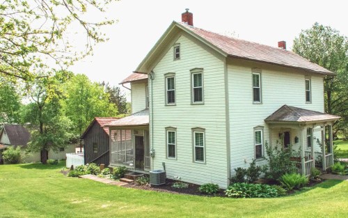 A charming two-story white house with a porch, surrounded by green grass and trees on a sunny day.