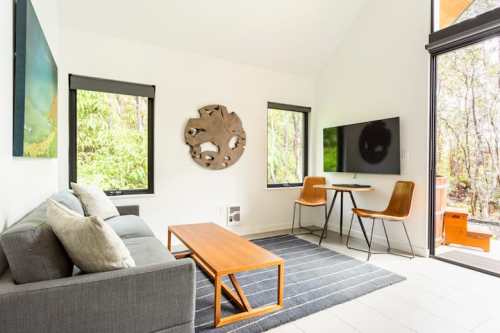 A modern living space with a gray sofa, wooden coffee table, and a small dining area, surrounded by large windows.