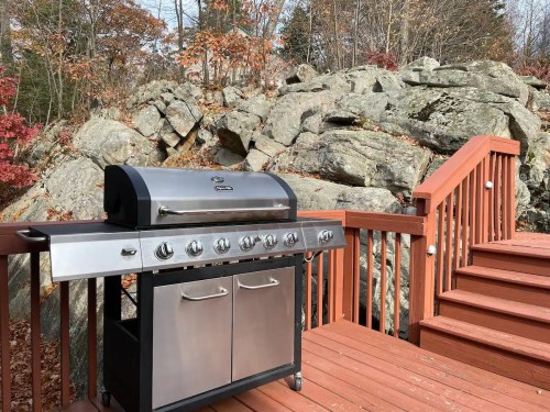 A stainless steel gas grill on a wooden deck, with rocky terrain and autumn foliage in the background.