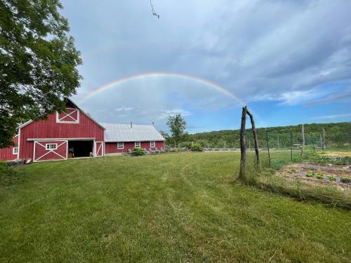 A red barn with a rainbow arching over a green field and garden under a cloudy sky.