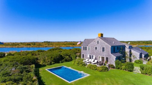 Aerial view of a modern house with a pool, surrounded by greenery and a river under a clear blue sky.