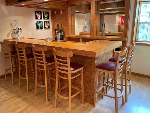 A wooden home bar with high stools, mirrors, and framed artwork on the walls, illuminated by natural light.