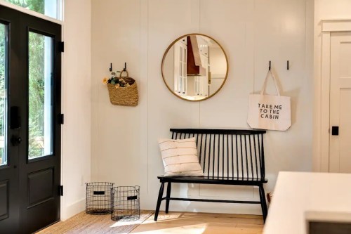 A cozy entryway featuring a black bench, round mirror, wall hooks, and a tote bag hanging beside decorative baskets.