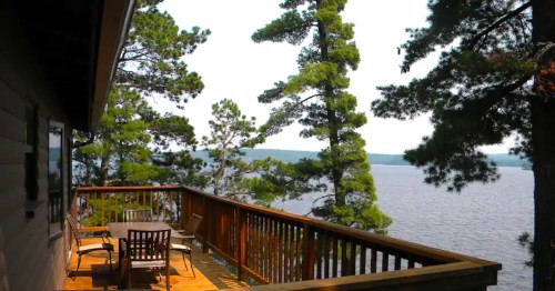 A wooden deck overlooking a serene lake, surrounded by tall pine trees under a clear sky.