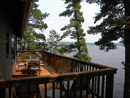 A wooden deck overlooking a serene lake, surrounded by tall pine trees under a clear sky.