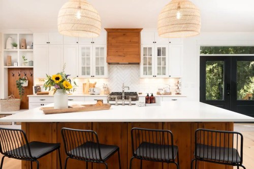 Bright kitchen with white cabinets, a large island, black chairs, and pendant lights, featuring a vase of sunflowers.