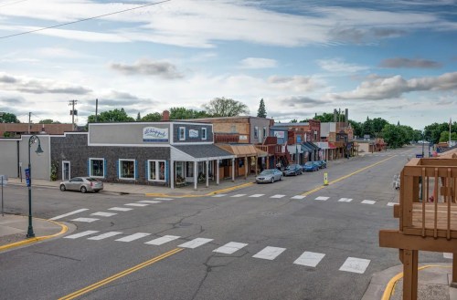 A quiet small town street with shops, parked cars, and a cloudy sky overhead.