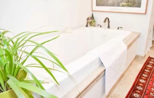 A modern bathroom featuring a white bathtub, a towel draped over the edge, and a potted plant beside a decorative rug.