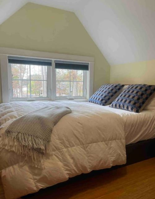 Cozy bedroom with a large bed, two patterned pillows, and natural light from windows. Soft blanket draped over the bed.