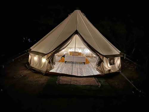 A cozy glamping tent at night, illuminated from within, featuring a bed and decorative rugs.