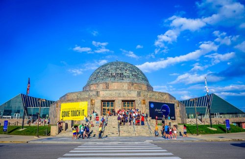 A large museum with a dome, featuring crowds of people outside under a blue sky with scattered clouds.