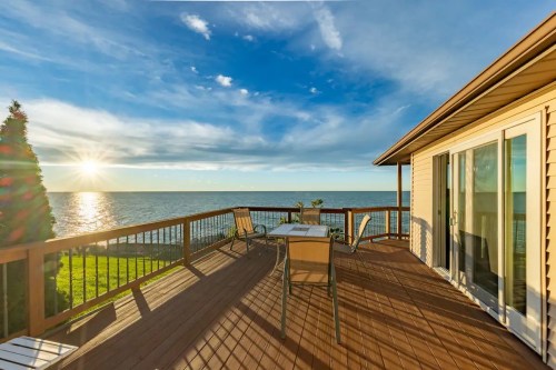 A sunlit deck overlooking a calm lake, with chairs and a table, surrounded by greenery and a clear blue sky.