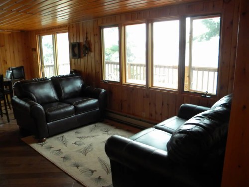 Cozy living room with two black leather sofas, wooden walls, and large windows overlooking a deck.