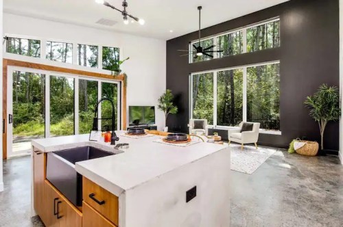 Modern kitchen with a large island, sleek appliances, and a spacious living area featuring large windows and greenery outside.