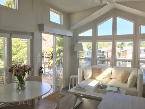 Bright, airy living space with large windows, a white sofa, dining table, and a view of a waterfront outside.