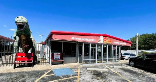 A barbecue restaurant with a large dinosaur statue outside, featuring a red awning and glass doors.